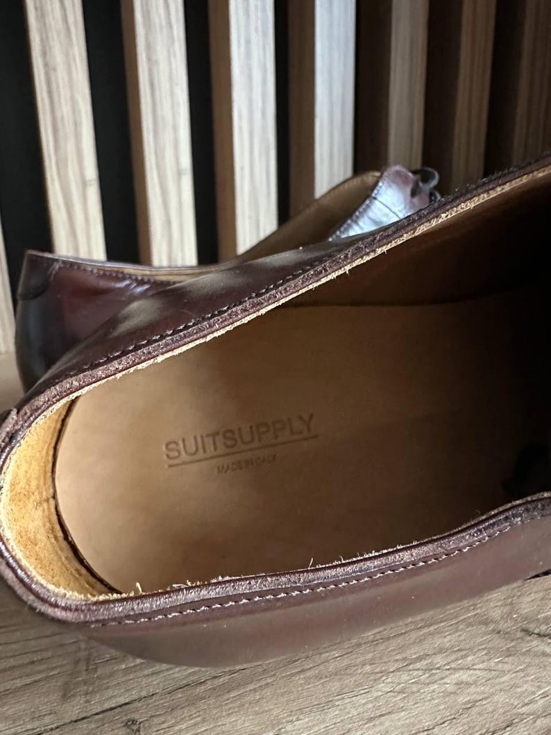 May include: Close-up of a pair of brown leather dress shoes. The interior of the shoe is visible, with the brand name "SUITSUPPLY" and "MADE IN ITALY" stamped on the insole. The shoes have a classic design and are made of high-quality leather.