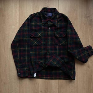 May include: A long-sleeved plaid shirt in a dark color palette of green, red, and yellow. The shirt has two chest pockets and a button-down front. The collar is a classic style, and the cuffs are buttoned.