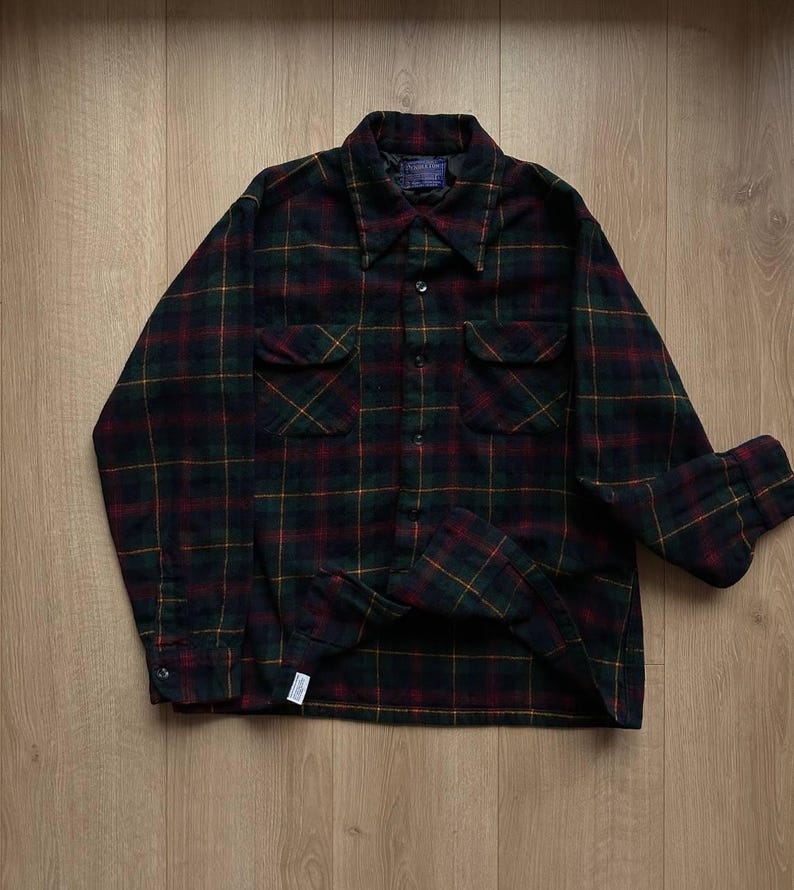 May include: A long-sleeved plaid shirt in a dark color palette of green, red, and yellow. The shirt has two chest pockets and a button-down front. The collar is a classic style, and the cuffs are buttoned.