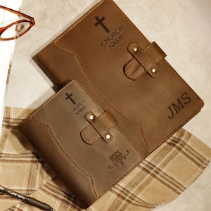 May include: Two brown leather journals with cross and text engravings. The larger journal has "CHURCH NAME" and "JMS" engraved. The smaller journal has "CHURCH NAME" and "ROSS" engraved. Both have a strap closure.