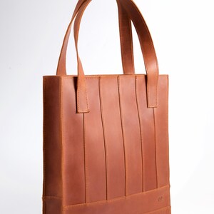 May include: A brown leather tote bag with vertical stitched details and two top handles. The bag has a rectangular shape and a structured design. The bag is made of a light brown leather material.