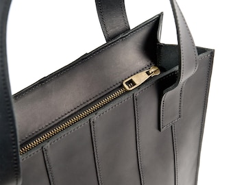 May include: Black leather tote bag with a vertical stitched design. The bag features a brass zipper closure and two top handles. The bag is made of a durable material and is suitable for everyday use.