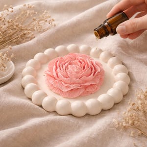 May include: A pink, flower-shaped soap sits on a white, beaded ceramic dish. A small brown bottle is held above the dish, with a drop of liquid falling. The dish is on a beige cloth, with dried flowers in the background.