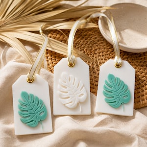 May include: Three rectangular white wax tags, each featuring a raised monstera leaf design. Two tags display a mint green leaf, while the centre tag features a white leaf. Each tag is suspended by a white ribbon with gold trim through a gold eyelet.