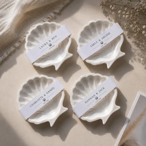 May include: Four white ceramic shell-shaped dishes, each wrapped with a white paper band. The bands feature printed names: Laura & Ben, Emily & Archie, Charlotte & Daniel, and Hannah & Jack, along with the date 16.05.2026 and a small shell icon.