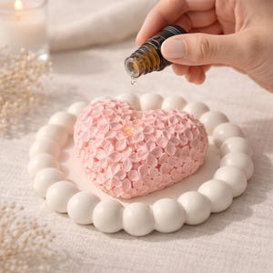 May include: A pink heart-shaped ornament with a floral pattern, on a white plate with a beaded border. A small brown bottle is held above the heart, with a drop of liquid falling onto it.