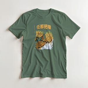 Può includere: T-shirt verde con un design grafico con testo giapponese e un'illustrazione astratta in giallo, marrone e bianco. Il design sembra essere una rappresentazione stilizzata di una creatura o di un oggetto.