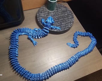May include: A blue articulated lizard toy, 3D printed, lies on a wooden surface. The lizard is in a heart shape, with a separate tail piece. A small, round, brick-patterned base and a dark object are in the background.