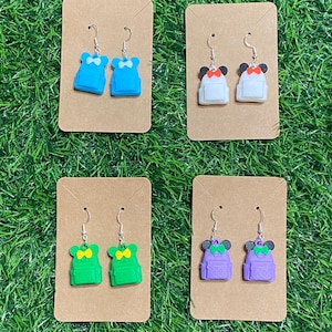 Op de afbeelding: Vier paar fantasie-oorbellen, elk met een rugzakontwerp. De oorbellen zijn blauw, wit, groen en paars, met strikken en muisoren details. Ze hangen aan zilveren haken en worden gepresenteerd op bruin karton.