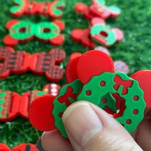 May include: A collection of small, red and green plastic accessories shaped like mouse ears, featuring various holiday-themed designs including wreaths with bows, plaid patterns, and Santa suit motifs with yellow belt buckles, arranged in rows.