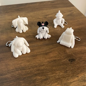 May include: A collection of five small animal and character keychains. Four are solid white figures including an elephant, a monkey, a robot, and a long-eared creature. The fifth is a white dog wearing a black hat with a small red heart detail.