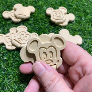 May include: A collection of beige, 3D-printed plastic clips shaped like the head of a cartoon mouse. Each clip features a raised outline of the character's face, including rounded ears, eyes, and a nose, with a hinged mechanism on the bottom section.