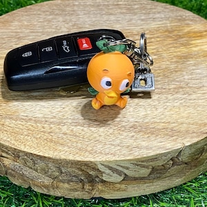 May include: A 3D-printed orange bird keychain with a key fob. The bird has a round orange body, green leaves, and a yellow beak. The black key fob has buttons for locking and unlocking a vehicle.