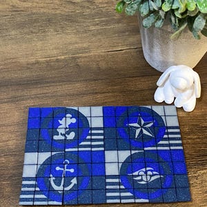 May include: A blue and gray mosaic coaster featuring nautical and Disney-themed designs. The coaster has a grid pattern with images of an anchor, a star, and a Mickey Mouse silhouette. A small white dog figurine and a potted plant are in the background.
