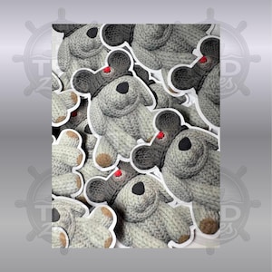 May include: A collection of multiple identical stuffed animal toys made of light gray knit fabric. Each toy features a dark gray knit hat with a small red pom-pom on top, a black nose, and tan-colored feet. The toys are arranged in a layered pattern.