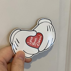 Puede incluir: Una pegatina de guante estilo dibujo animado en blanco con contornos negros, que muestra dos manos juntas sosteniendo un corazón rojo. El corazón contiene el texto blanco "LOVE YOUR DOOR!" en cursiva. La pegatina tiene un acabado brillante.