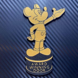 Könnte beinhalten: Eine goldfarbene Mickey Mouse-Figur in einem Trophäendesign. Die Figur steht auf einem schwarzen Sockel mit den Worten "AWARD WINNING DOOR" in goldfarbenen Buchstaben. Der Hintergrund ist eine dunkelblaue, strukturierte Oberfläche.