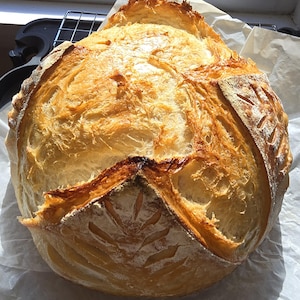 May include: A freshly baked loaf of artisan bread with a golden-brown crust. The bread has intricate scoring patterns and a rustic appearance, resting on parchment paper. The crust is crispy, and the interior appears soft and airy.