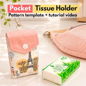 Peut inclure: Un étui à mouchoirs de poche rectangulaire en tissu avec un rabat rose et un corps blanc orné d'un motif sur le thème de Paris, incluant la tour Eiffel. À côté se trouvent un paquet de mouchoirs en bambou et une sacoche banane rayée rose et blanc.