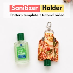 May include: A fabric hand sanitizer holder with a floral pattern in orange, yellow, and cream, featuring a metal lobster clasp and a red snap button. Next to it is a clear plastic bottle of green hand sanitizer. Text reads: Sanitizer Holder.