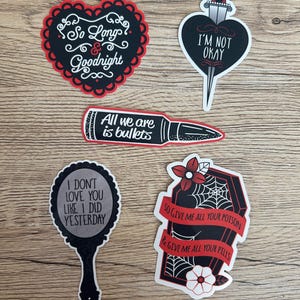 May include: A collection of stickers with gothic and romantic themes. Designs include a heart with a dagger, a heart-shaped sticker with the text "I'm Not Okay", a bullet, a mirror, and a coffin with text and spiderwebs.