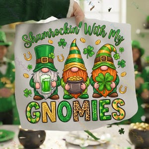 May include: A St. Patrick's Day themed design featuring three gnomes with green hats and beards, holding a beer, a pot of gold, and a four-leaf clover. The text "Shamrockin' With My Gnomies" is displayed in green and gold.