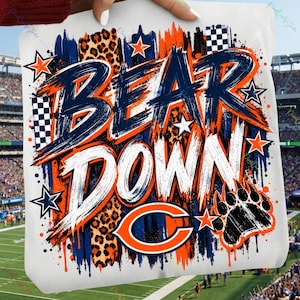 May include: A white t-shirt featuring a graphic design with the text BEAR DOWN in navy blue and white brushstroke lettering. The design includes orange and navy blue paint splatters, leopard print accents, stars, and a bear paw print.