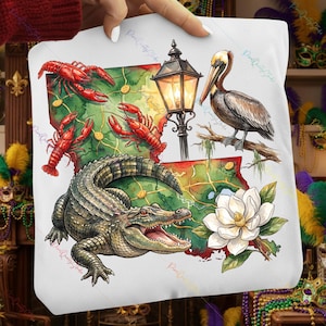 Puede incluir: Bolso tote blanco con un diseño de acuarela de Luisiana. El diseño incluye cangrejos rojos, un caimán, un pelícano, una flor de magnolia y una farola. El fondo es un mapa de Luisiana.