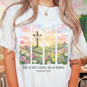 Puede incluir: Camiseta blanca con un diseño de acuarela de una cruz sobre una colina con un amanecer, enmarcada por líneas verticales. El diseño incluye flores coloridas y el texto "He is not here; He is Risen. Matthew 28:6."
