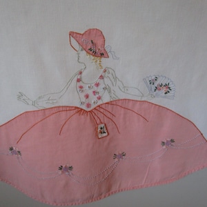 May include: Embroidered tablecloth featuring a pink and white design of a woman in a pink dress and hat holding a fan. The tablecloth has a scalloped edge.