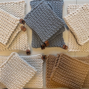 Puede incluir: Un conjunto de agarraderas y salvamanteles cuadrados tejidos en colores neutros como crema, gris y marrón. Cada juego tiene una etiqueta de cuero. El tejido grueso sugiere resistencia al calor.
