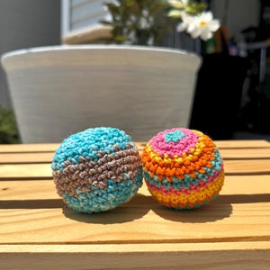 Puede incluir: Dos bolas esf&eacute;ricas de ganchillo. La bola de la izquierda presenta un patr&oacute;n variado de hilo azul claro, turquesa y marr&oacute;n. La bola de la derecha muestra rayas horizontales de hilo rosa, naranja, amarillo y azul claro en punto circular.