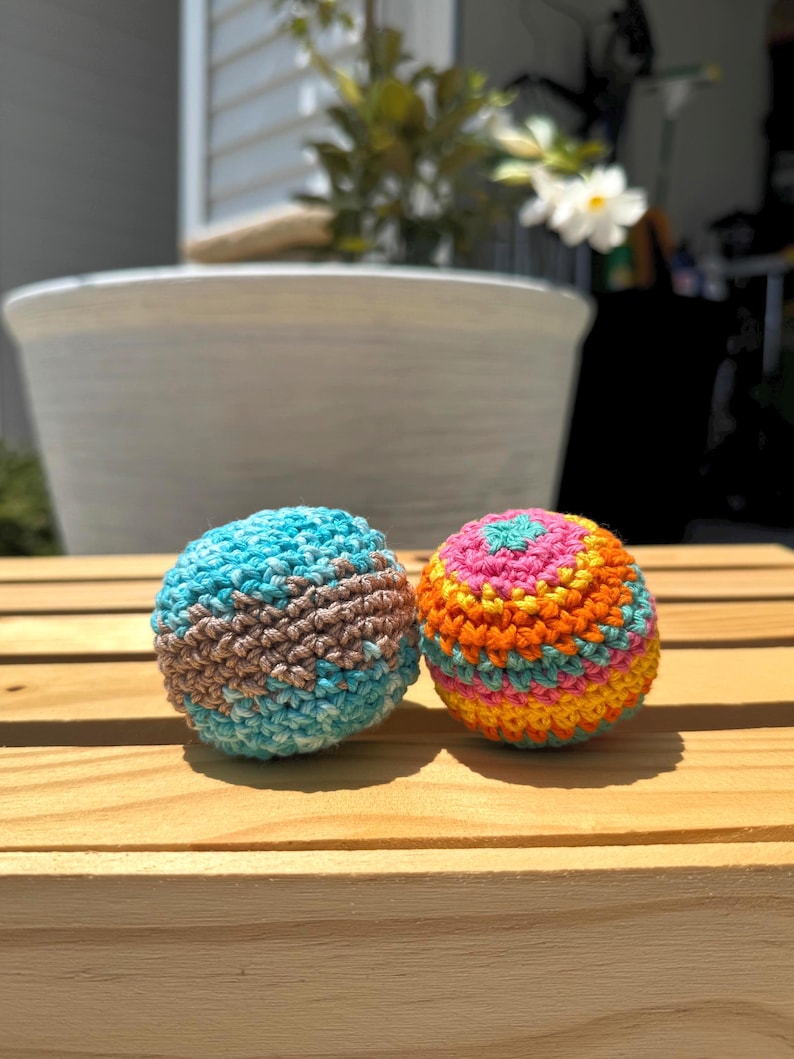 Puede incluir: Dos bolas esf&eacute;ricas de ganchillo. La bola de la izquierda presenta un patr&oacute;n variado de hilo azul claro, turquesa y marr&oacute;n. La bola de la derecha muestra rayas horizontales de hilo rosa, naranja, amarillo y azul claro en punto circular.