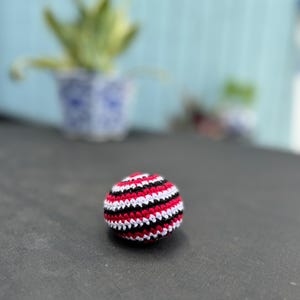 Puede incluir: Una peque&ntilde;a pelota de ganchillo hecha a mano, de forma esf&eacute;rica, con un patr&oacute;n repetitivo de rayas horizontales en hilo rojo vibrante, blanco n&iacute;tido y negro s&oacute;lido. La superficie texturizada est&aacute; formada por puntos apretados.