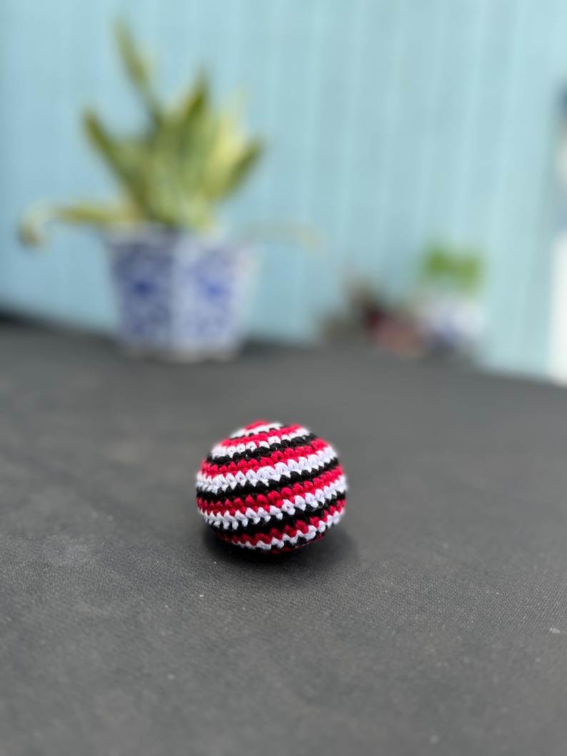 Puede incluir: Una peque&ntilde;a pelota de ganchillo hecha a mano, de forma esf&eacute;rica, con un patr&oacute;n repetitivo de rayas horizontales en hilo rojo vibrante, blanco n&iacute;tido y negro s&oacute;lido. La superficie texturizada est&aacute; formada por puntos apretados.