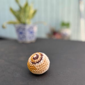 Puede incluir: Un peque&ntilde;o objeto esf&eacute;rico de ganchillo que presenta un patr&oacute;n de remolinos de hilo en tonos crema, bronceado, marr&oacute;n claro y marr&oacute;n oscuro. La textura est&aacute; creada por puntos apretados y uniformes que forman una forma redondeada y densa.