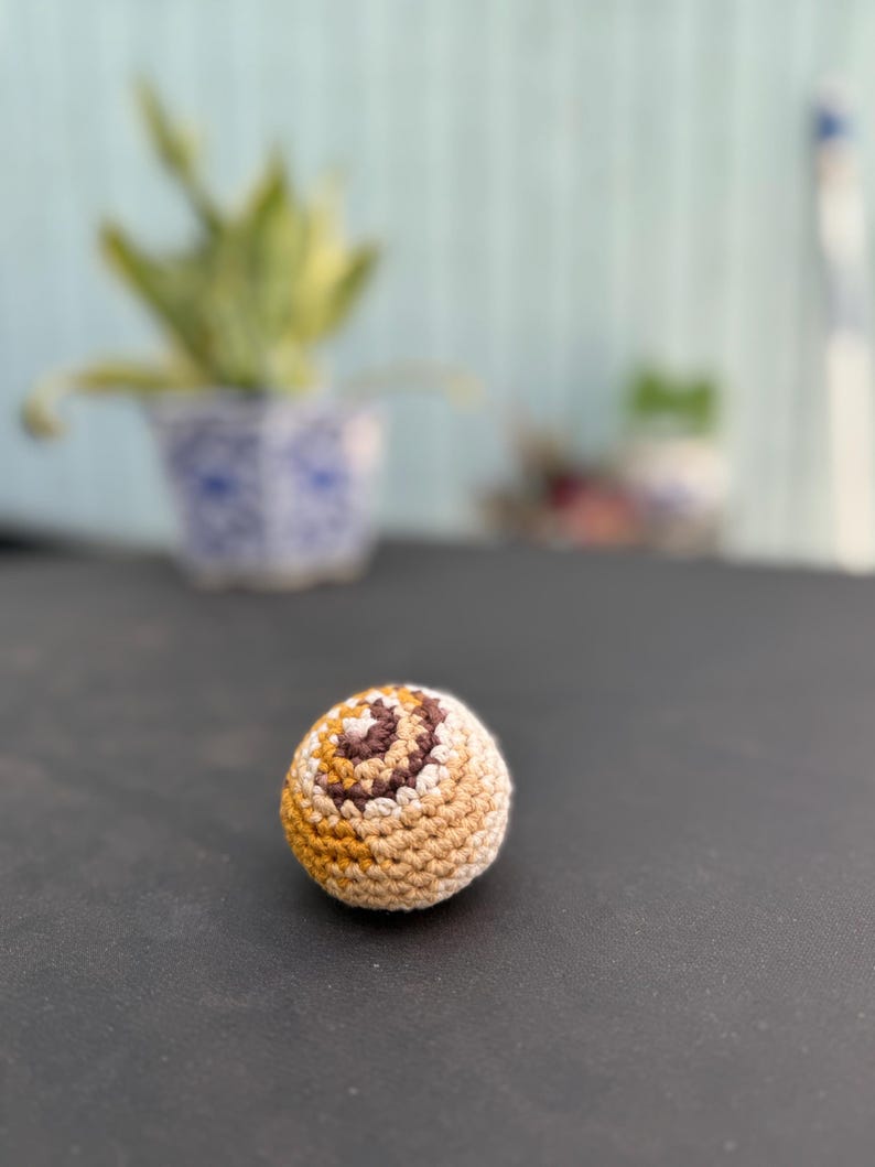 Puede incluir: Un peque&ntilde;o objeto esf&eacute;rico de ganchillo que presenta un patr&oacute;n de remolinos de hilo en tonos crema, bronceado, marr&oacute;n claro y marr&oacute;n oscuro. La textura est&aacute; creada por puntos apretados y uniformes que forman una forma redondeada y densa.