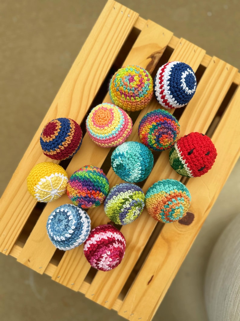 Puede incluir: Una colecci&oacute;n de trece peque&ntilde;as bolas redondas de ganchillo con varios patrones coloridos, incluyendo tonos brillantes s&oacute;lidos, hilo multicolor jaspeado, un dise&ntilde;o de sand&iacute;a con semillas negras y un motivo de rodaja de c&iacute;trico en amarillo y blanco.