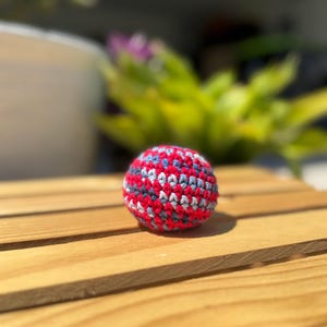 Puede incluir: Una peque&ntilde;a pelota de ganchillo hecha a mano, de forma esf&eacute;rica, con un patr&oacute;n variado de hilo rojo brillante, gris carb&oacute;n oscuro y azul claro. La superficie texturizada est&aacute; creada con puntos apretados y uniformes, formando un objeto denso.