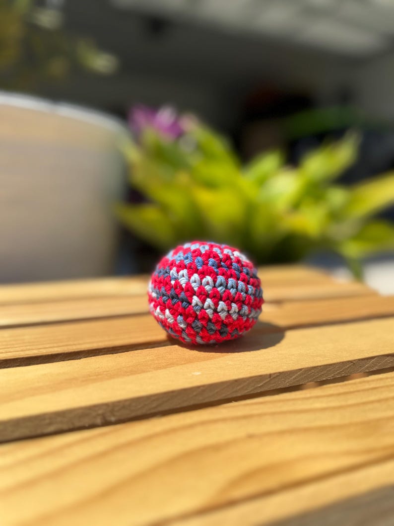 Puede incluir: Una peque&ntilde;a pelota de ganchillo hecha a mano, de forma esf&eacute;rica, con un patr&oacute;n variado de hilo rojo brillante, gris carb&oacute;n oscuro y azul claro. La superficie texturizada est&aacute; creada con puntos apretados y uniformes, formando un objeto denso.