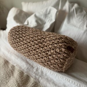 Puede incluir: Un cojín cilíndrico de ganchillo marrón claro con una superficie texturizada. El cojín descansa sobre una cama blanca con almohadas blancas en el fondo. Una pequeña etiqueta marrón es visible en el lateral del cojín.