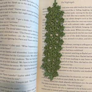 Puede incluir: Un marcapáginas de ganchillo verde hecho a mano con un diseño de hojas, colocado en un libro abierto. El marcapáginas está hecho de un hilo suave y texturizado y añade un toque de elegancia a cualquier experiencia de lectura.