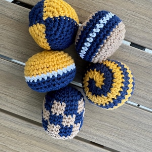 Puede incluir: Cinco bolas redondas de ganchillo con varios patrones y combinaciones de colores. Una bola tiene un patr&oacute;n de bloques amarillo y azul marino, otra un dise&ntilde;o de rayas azul marino y beige con un centro blanco, y una presenta una espiral amarilla y azul marino.