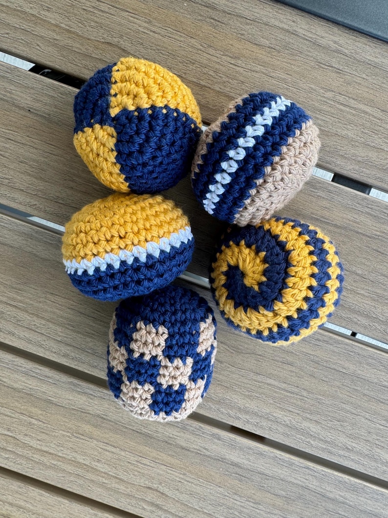 Puede incluir: Cinco bolas redondas de ganchillo con varios patrones y combinaciones de colores. Una bola tiene un patr&oacute;n de bloques amarillo y azul marino, otra un dise&ntilde;o de rayas azul marino y beige con un centro blanco, y una presenta una espiral amarilla y azul marino.
