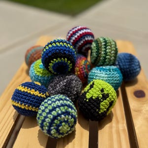 Puede incluir: Una colecci&oacute;n de peque&ntilde;as bolas redondas de ganchillo en varios colores y patrones, incluyendo azul, verde, negro, rojo y blanco. Cada bola presenta un dise&ntilde;o de hilo texturizado distinto, con algunas mostrando rayas, espirales o bloques.
