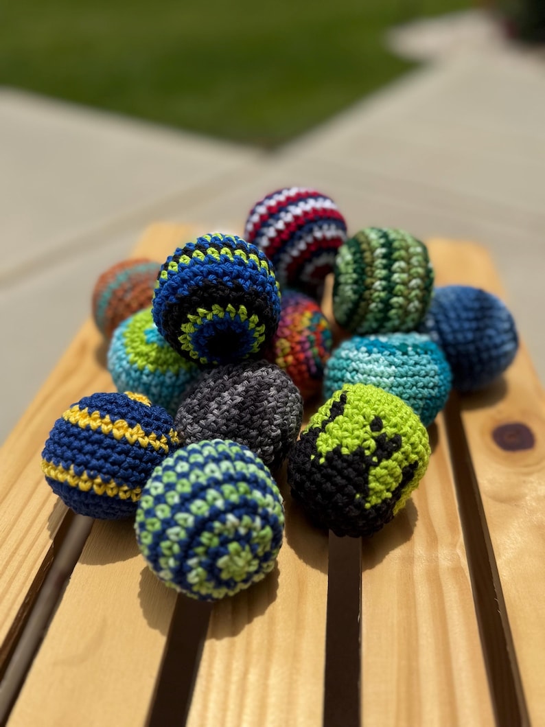 Puede incluir: Una colecci&oacute;n de peque&ntilde;as bolas redondas de ganchillo en varios colores y patrones, incluyendo azul, verde, negro, rojo y blanco. Cada bola presenta un dise&ntilde;o de hilo texturizado distinto, con algunas mostrando rayas, espirales o bloques.