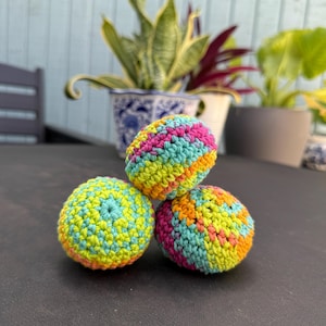 Puede incluir: Tres objetos esf&eacute;ricos elaborados con hilo multicolor mediante una t&eacute;cnica de ganchillo. El hilo presenta una mezcla vibrante de rosa brillante, verde lima, turquesa y naranja, creando un patr&oacute;n texturizado y variado en cada superficie.