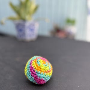 Puede incluir: Una peque&ntilde;a pelota esf&eacute;rica tejida a mano con un vibrante patr&oacute;n de hilo multicolor. La superficie muestra una mezcla arremolinada de puntos en rosa brillante, amarillo sol, naranja y azul turquesa claro, creando una apariencia suave y texturizada.