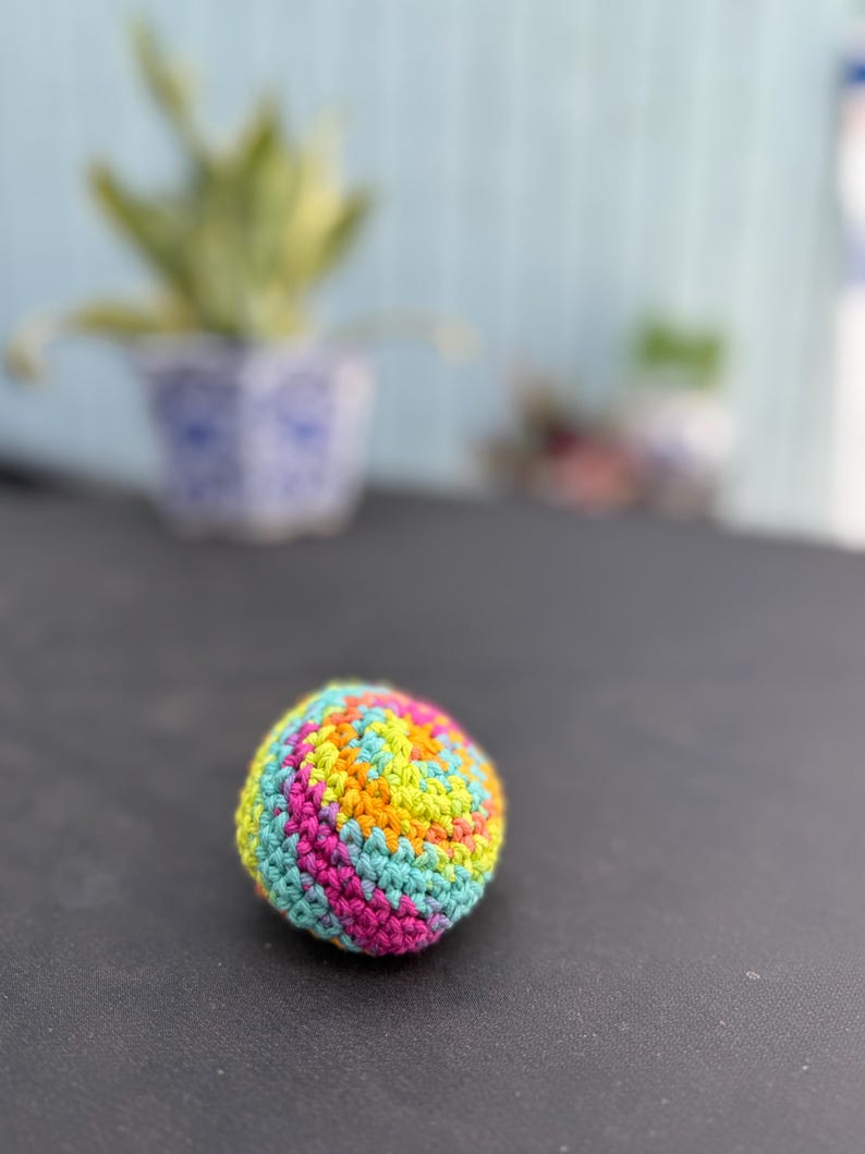 Puede incluir: Una peque&ntilde;a pelota esf&eacute;rica tejida a mano con un vibrante patr&oacute;n de hilo multicolor. La superficie muestra una mezcla arremolinada de puntos en rosa brillante, amarillo sol, naranja y azul turquesa claro, creando una apariencia suave y texturizada.