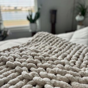 Puede incluir: Primer plano de una manta de punto grueso en un tono beige neutro. La textura de la manta es prominente, con hilos gruesos y en bucle que crean una apariencia suave y acogedora. La manta está extendida sobre una cama, lo que sugiere un ambiente acogedor y confortable.